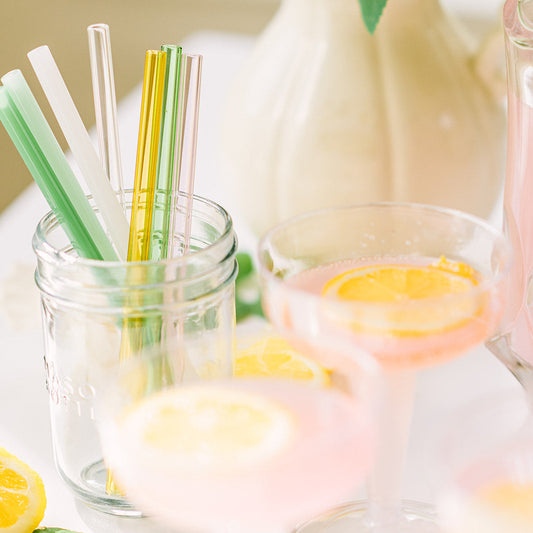 Colorful glass straws in a mason jar beside pink citrus cocktails
